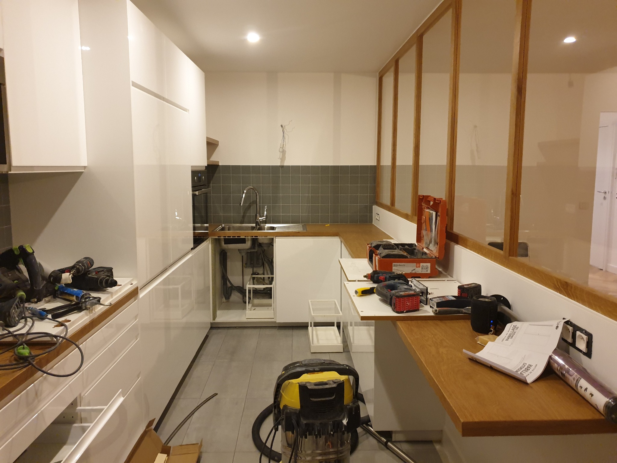 Wnętrze kuchni w trakcie montażu: białe szafki z drewnianymi blatami, widoczne narzędzia, odkurzacz przemysłowy i materiały wykończeniowe, szare płytki na ścianie nad zlewem, drewniana konstrukcja...