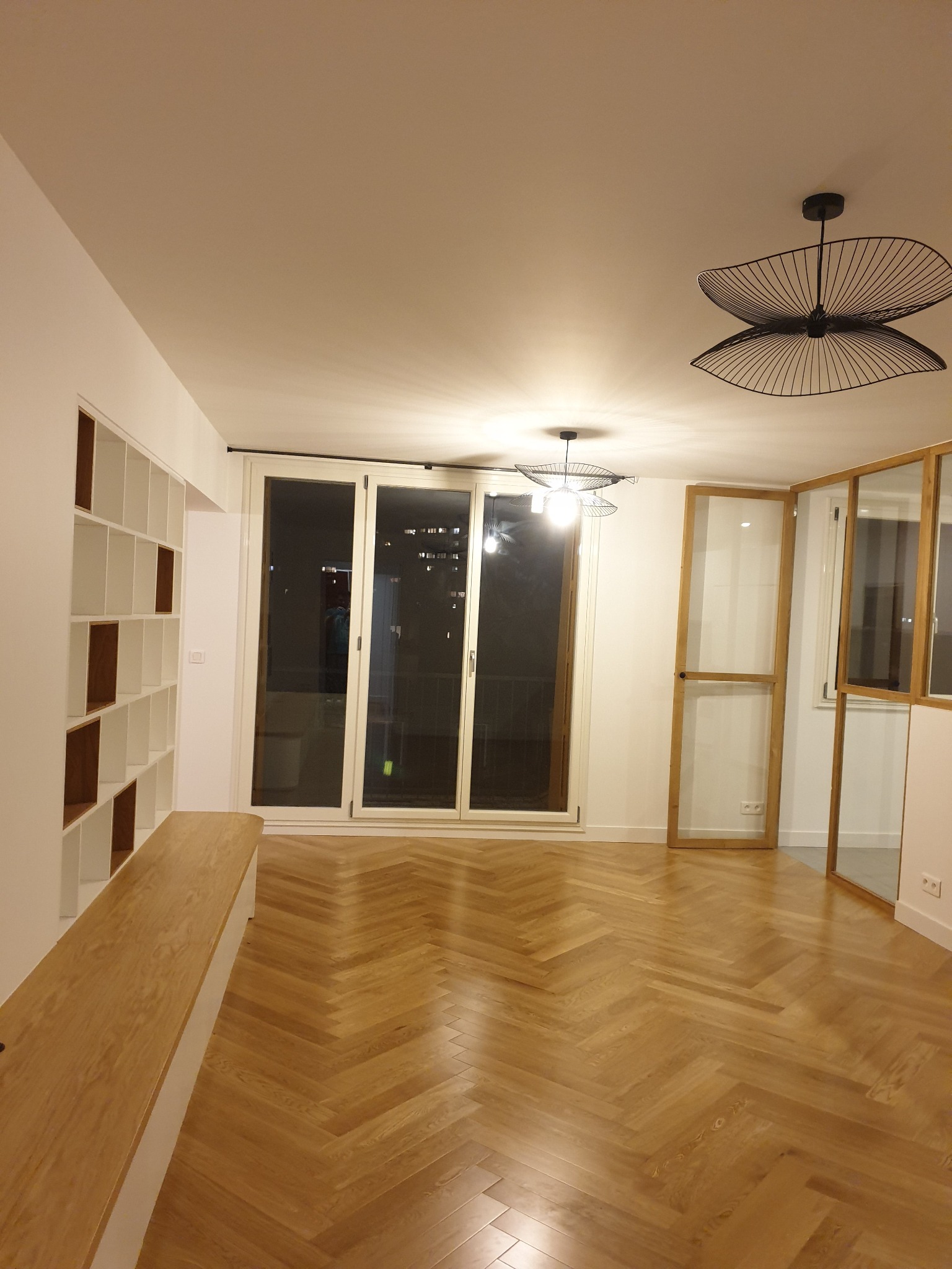 Jasne wnętrze po remoncie z parkietem ułożonym w jodełkę, zabudową regałową i nowoczesnym oświetleniem wiszącym.