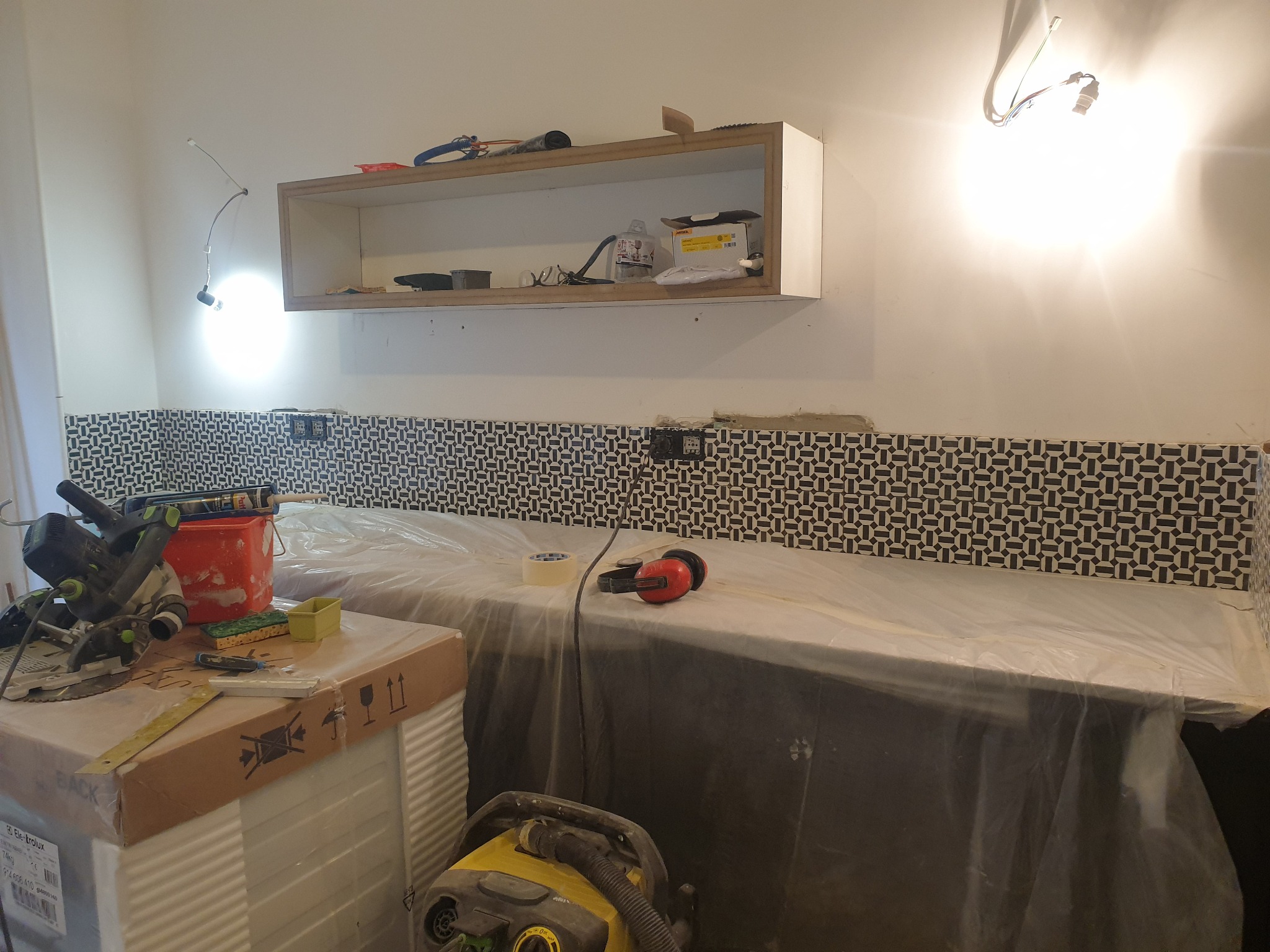 Fragment kuchni w trakcie układania dekoracyjnych płytek ściennych z geometrycznym wzorem; blat zabezpieczony folią, widoczne narzędzia, odkurzacz i górna szafka bez frontów.