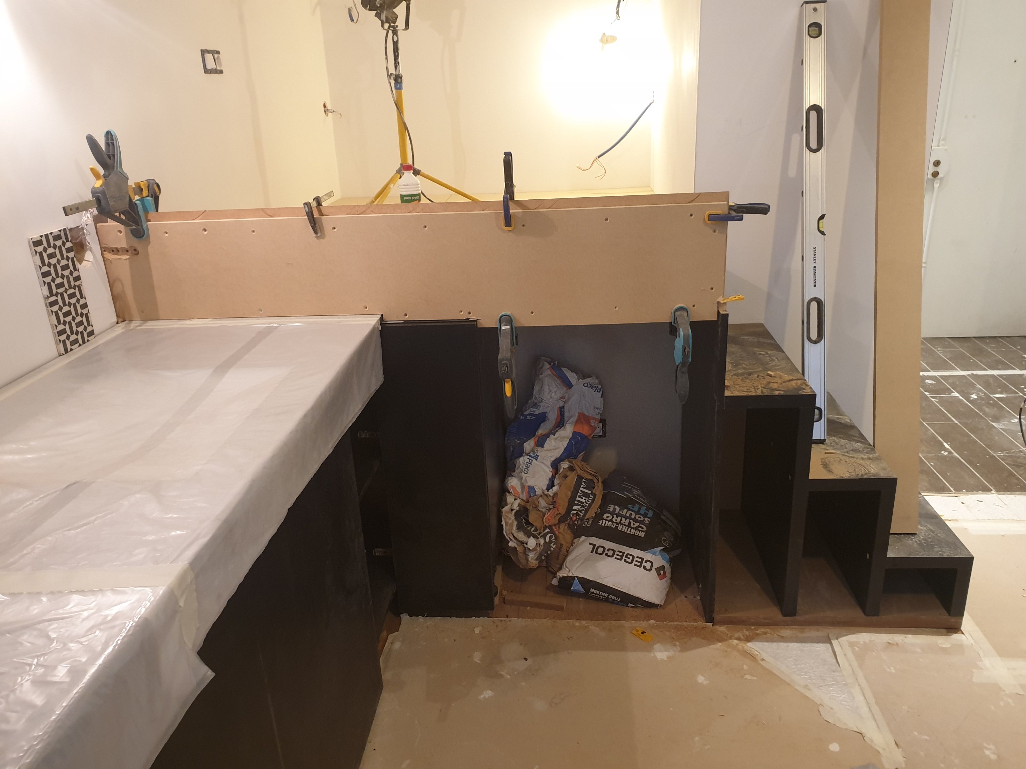 Montaż zabudowy kuchennej w trakcie realizacji: blat zakryty folią ochronną, przytwierdzona listwa MDF za pomocą ścisków stolarskich, widoczne worki z zaprawą klejową CEGECOL, poziomica Stanley...