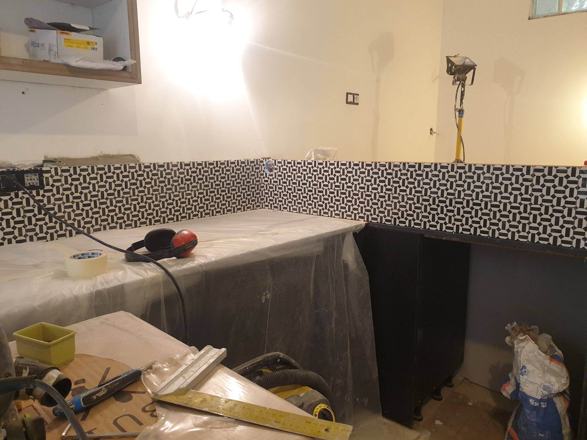 Układanie czarno-białej mozaiki na ścianie w kuchni, widoczne narzędzia pracy, słuchawki ochronne i zakryty folią blat.