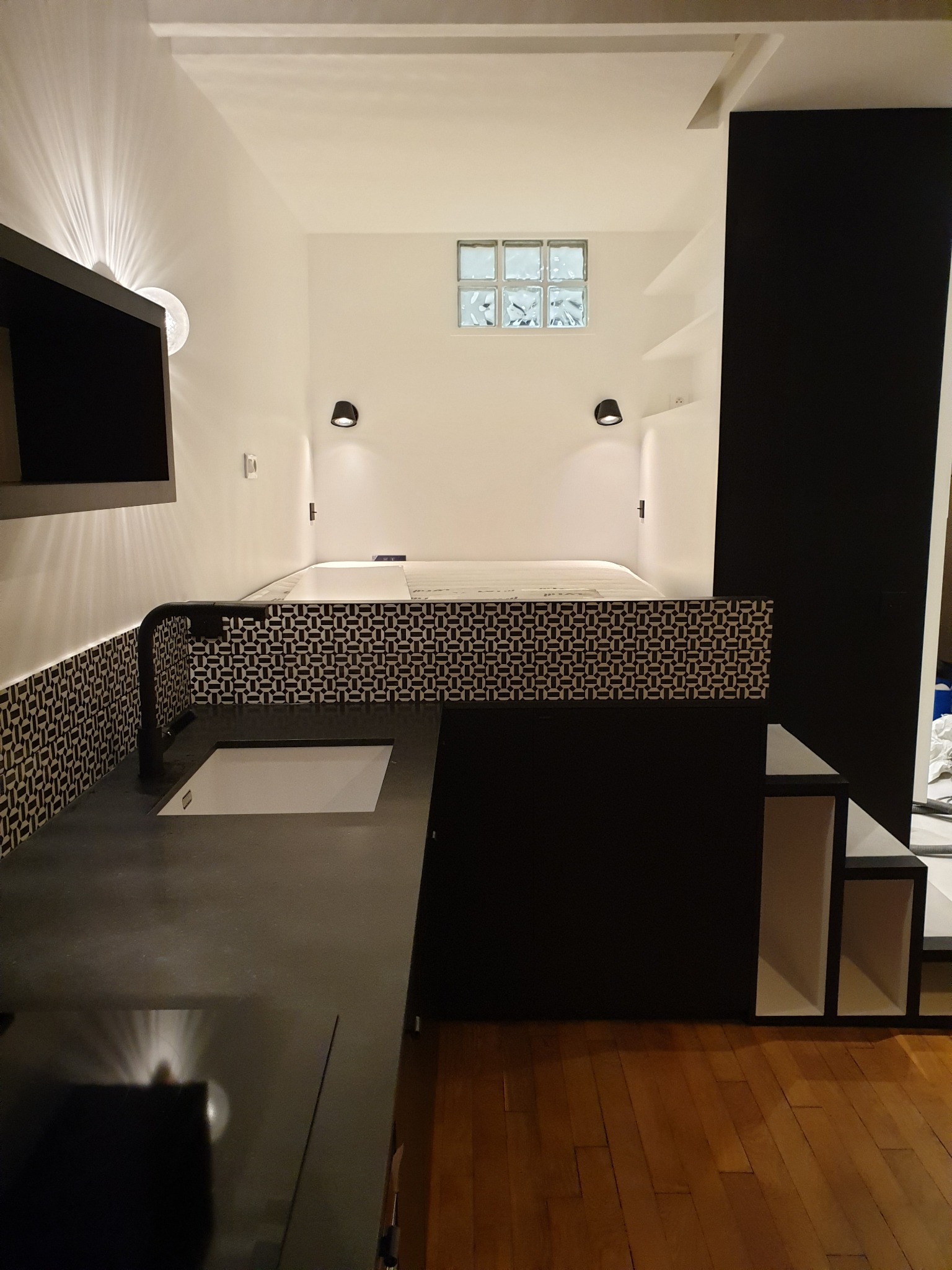 Kompaktowa, nowoczesna kawalerka z antresolą sypialną nad aneksem kuchennym; czarno-biała kolorystyka, wzorzysta mozaika na ścianie, drewniana podłoga, oświetlenie punktowe i szklane luksfery w oknie.