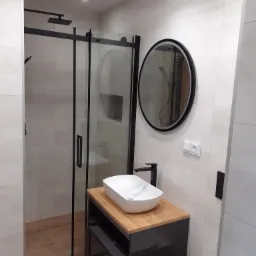 Nowoczesna łazienka z kabiną prysznicową typu walk-in z czarnymi ramami, okrągłym lustrem z czarną obwódką, białą umywalką nablatową na szafce z drewnianym blatem i czarnymi frontami oraz płytkami...