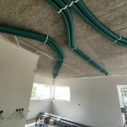 Zielone, karbowane rury wentylacyjne przymocowane do sufitu z płyty OSB za pomocą białych opasek zaciskowych w surowym stanie budynku, widoczne okna i narzędzia w tle.