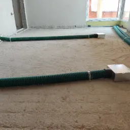 Zielone, karbowane rury wentylacyjne ułożone na podłodze w surowym stanie, z podłączonymi srebrnymi skrzynkami dystrybucyjnymi, widoczne okno z profilem Alufrof.