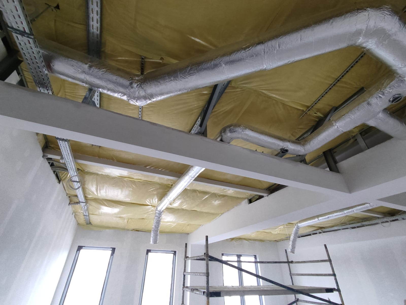 Instalacja przewodów wentylacyjnych owiniętych folią aluminiową pod sufitem ocieplonym żółtą wełną mineralną, widoczne elementy konstrukcji budynku i rusztowanie.