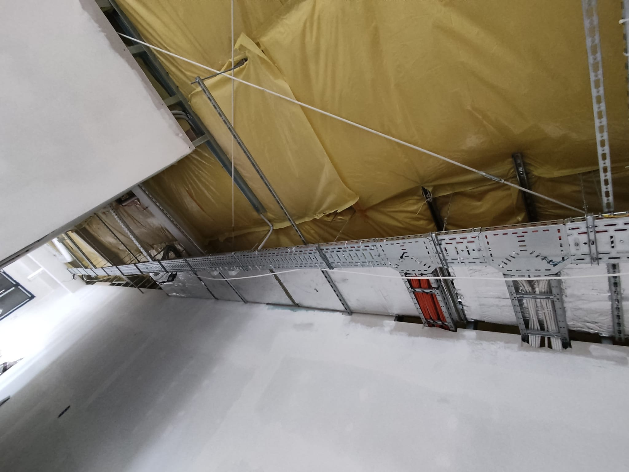 Widok z dołu na metalowe korytka kablowe zamontowane pod sufitem w surowym wnętrzu budynku, z widocznymi przewodami w kolorze pomarańczowym i białym, oraz izolacją termiczną w kolorze żółtym.