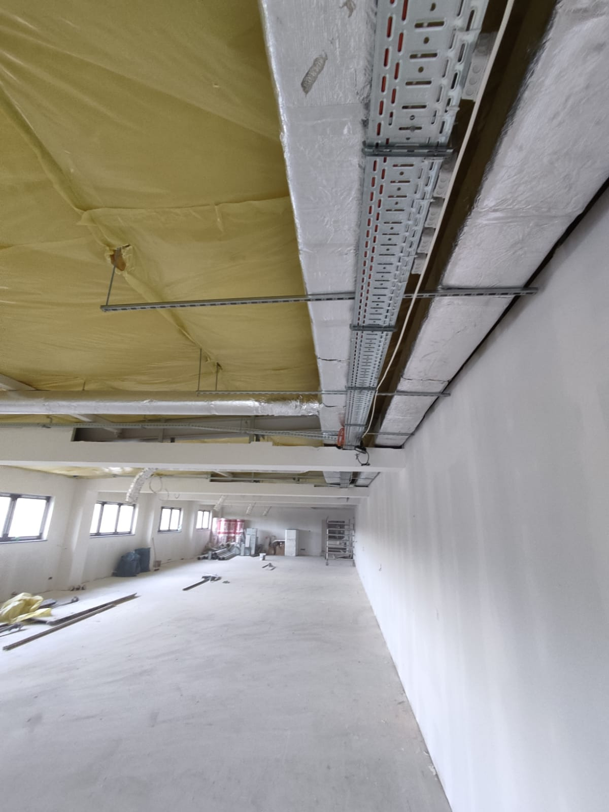 Wnętrze w trakcie montażu instalacji wentylacyjnej w budynku komercyjnym, widoczne koryta kablowe pod sufitem i izolacja termiczna, surowy stan wykończenia.