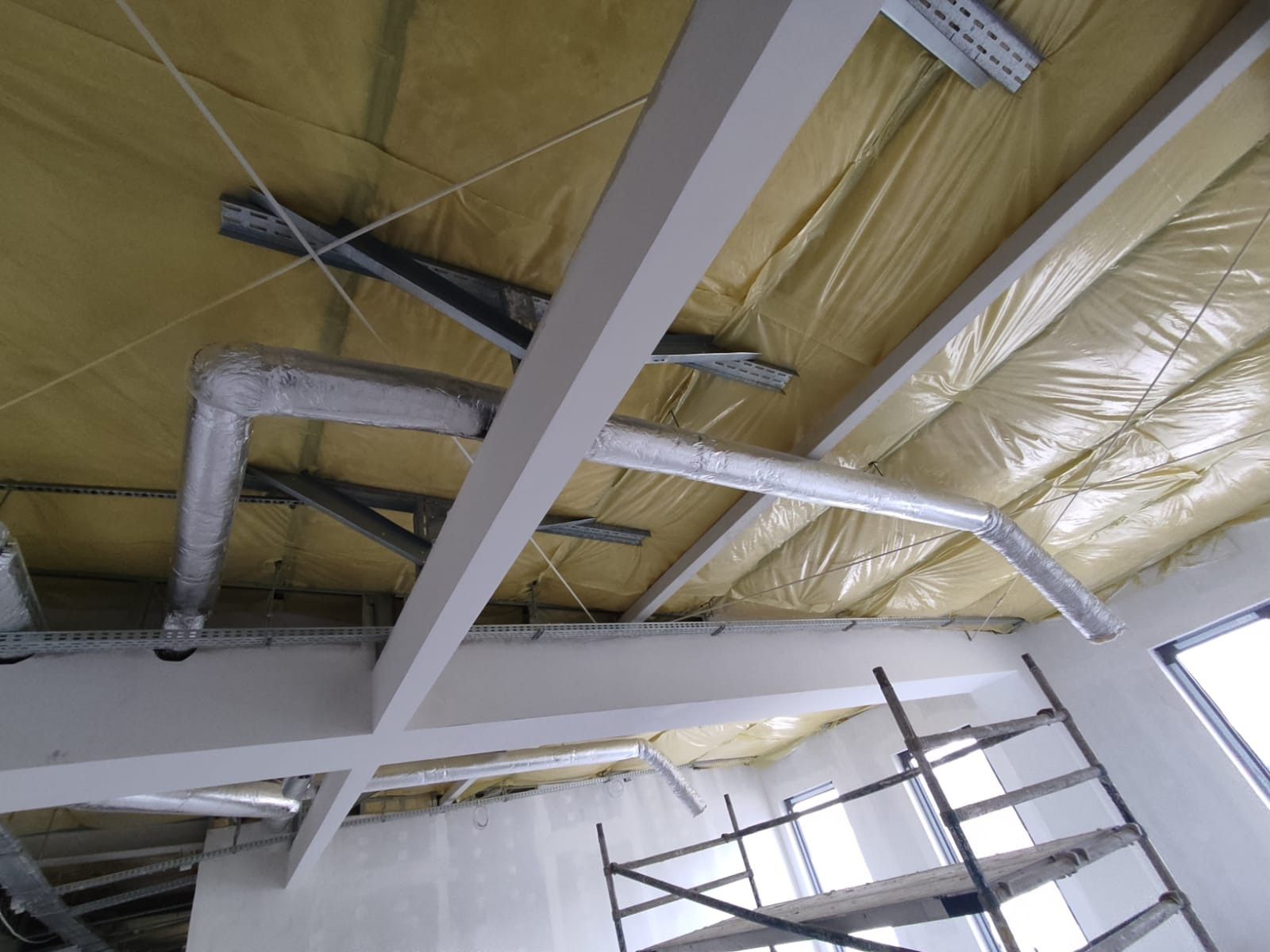 Wnętrze w trakcie prac instalacyjnych: widoczne ocieplenie stropu, metalowe konstrukcje podtrzymujące, aluminiowe przewody wentylacyjne oraz fragment rusztowania.