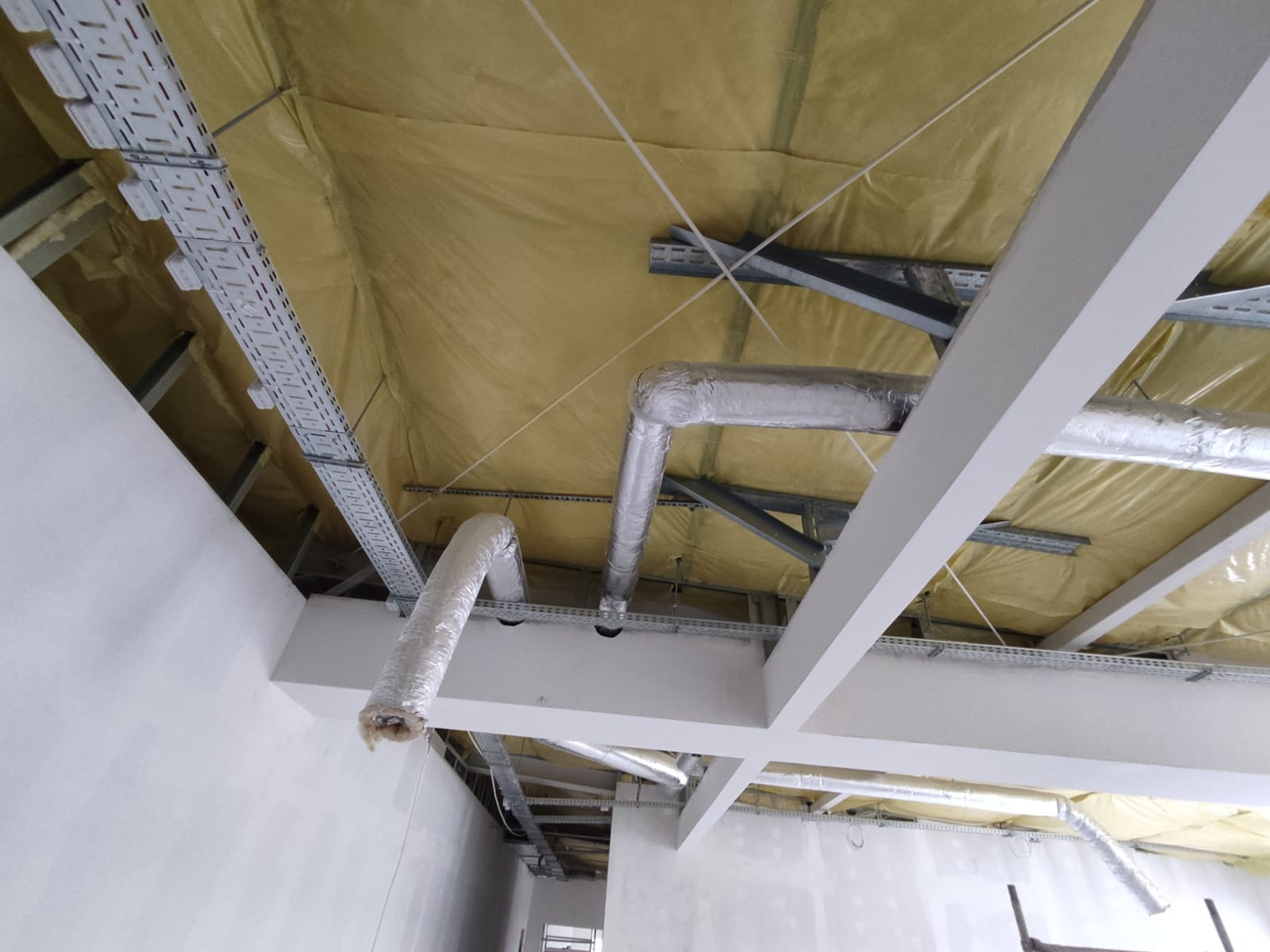 Montaż instalacji wentylacyjnej: widok na sufit z widocznymi kanałami wentylacyjnymi w izolacji, korytkami kablowymi oraz elementami konstrukcyjnymi podwieszanego sufitu pokrytego żółtą izolacją.