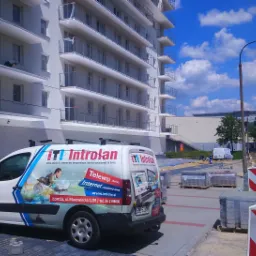 Biały van firmy Introlan z reklamą usług telekomunikacyjnych zaparkowany przed nowym budynkiem mieszkalnym w słoneczny dzień. Na karoserii widoczna grafika przedstawiająca osobę korzystającą...