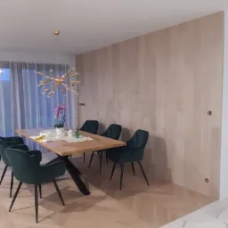 Jadalnia z drewnianą podłogą ułożoną we wzór jodełki, drewnianą ścianą z paneli, stołem z blatem z jasnego drewna i ciemnozielonymi krzesłami oraz nowoczesną lampą w kolorze złota.