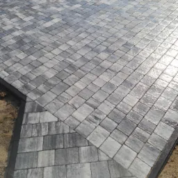 Świeżo ułożona, szara kostka brukowa z delikatnym połyskiem od wilgoci, ułożona w regularny wzór, z widoczną obwódką z ciemniejszej kostki, na granicy z piaszczystym podłożem.