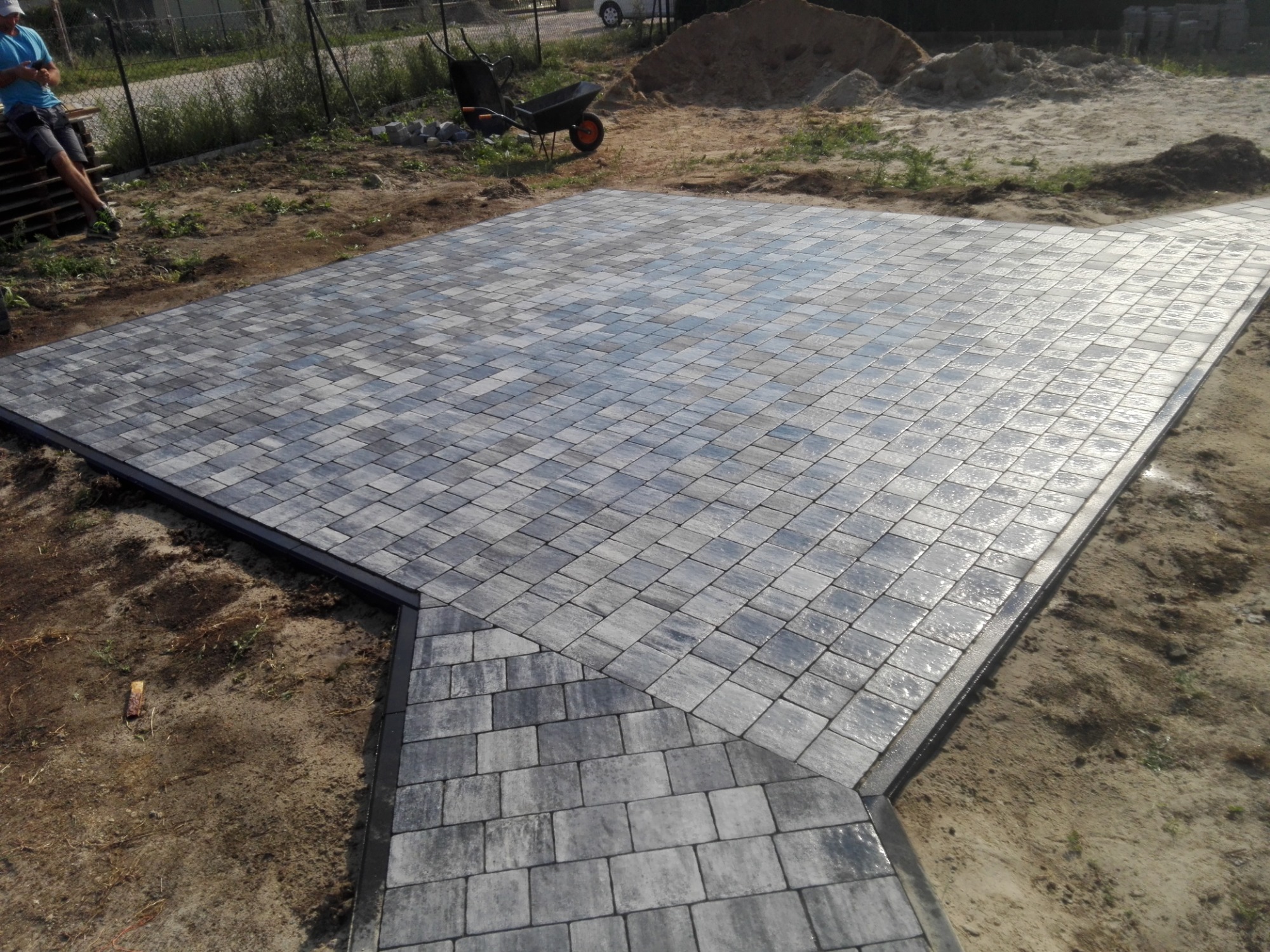 Nowo ułożona, szara kostka brukowa tworząca plac i ścieżkę w ogrodzie, z widoczną czarną obwódką i przygotowanym terenem pod dalsze prace.