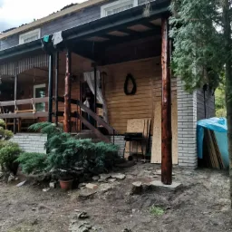 Drewniany dom z werandą w trakcie prac budowlanych, widoczne deski na dachu, materiały budowlane i narzędzia, teren wokół domu częściowo uporządkowany.