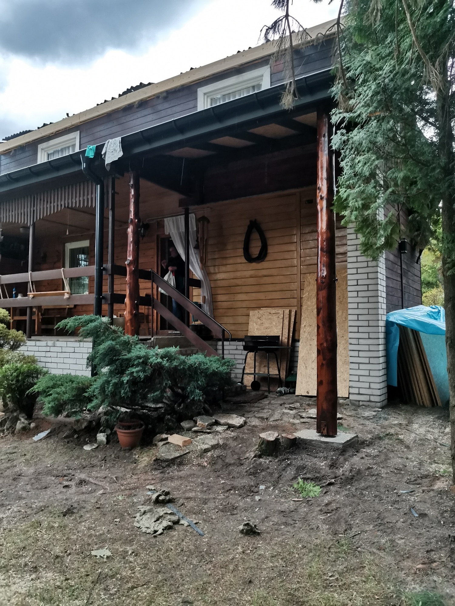 Drewniany dom z werandą w trakcie prac budowlanych, widoczne deski na dachu, materiały budowlane i narzędzia, teren wokół domu częściowo uporządkowany.