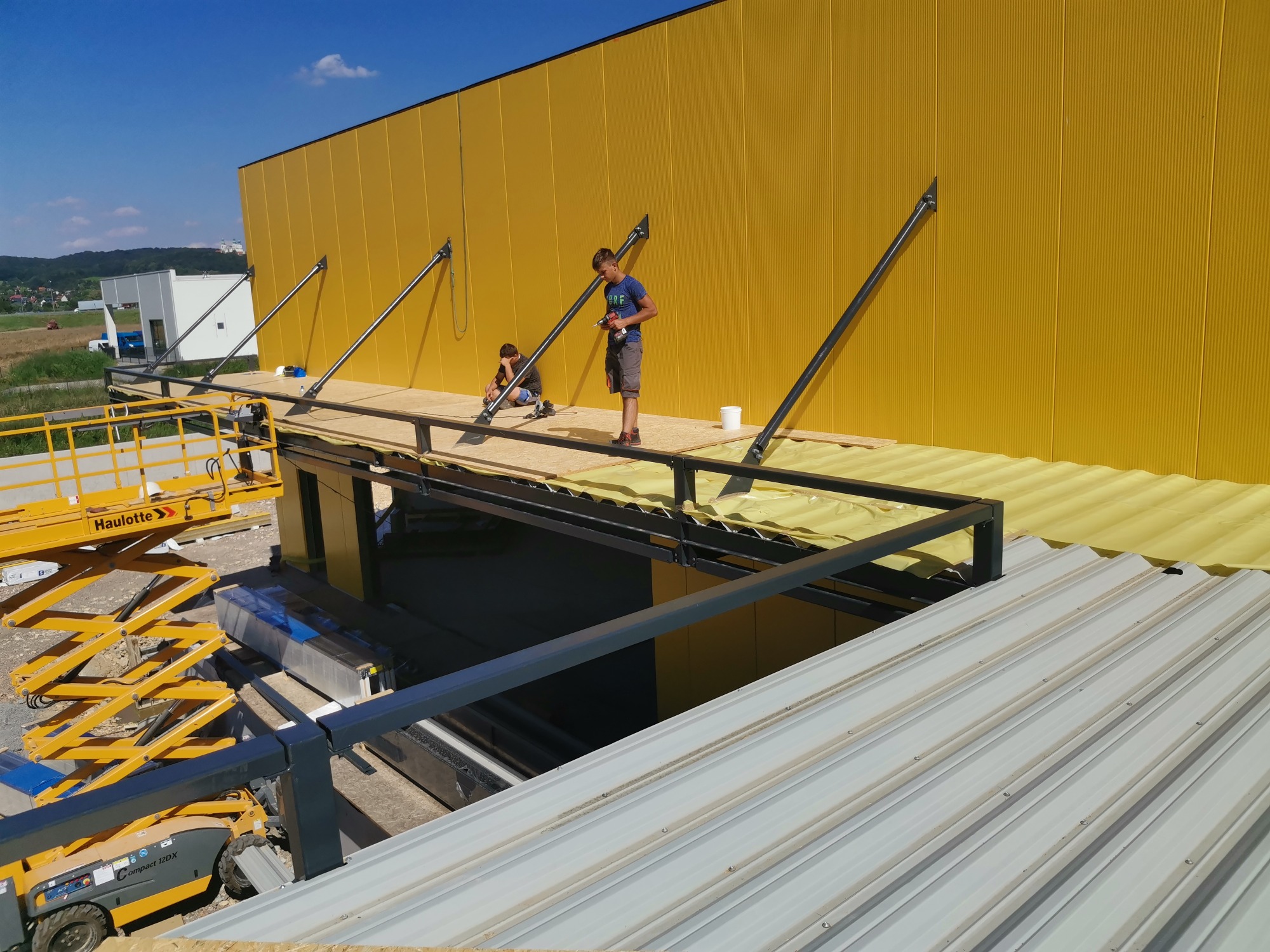 Dwóch monterów pracuje na platformie pod ścianą budynku o jaskrawożółtej elewacji, używając wiertarki i narzędzi, widoczny podnośnik koszowy Haulotte i fragment nowego dachu z blachy trapezowej.