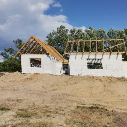 Wznoszenie ścian budynku z bloczków betonowych i drewnianej konstrukcji dachu w trakcie budowy, widoczne drzewa w tle.