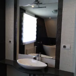 Nowoczesna łazienka z owalną umywalką nablatową, czarnym blatem i lustrem z oświetleniem, odbijającym wannę wolnostojącą i grzejnik drabinkowy.