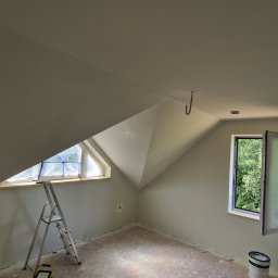AMJ SPERO SP. Z O.O. - Świeżo pomalowane poddasze ze skosami i oknami zabezpieczonymi folią. Widoczna drabina malarska i wiadro z farbą. Jasne, neutralne kolory ścian.