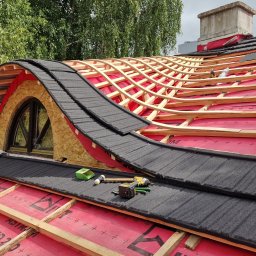 AMJ SPERO SP. Z O.O. - Konstrukcja dachu z oknem lukarny w trakcie krycia blachodachówką. Widoczne narzędzia i czerwona membrana dachowa. Dach z drewnianą więźbą i elementami poszycia.