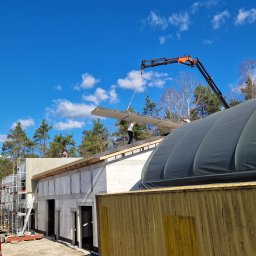 AMJ SPERO SP. Z O.O. - Montaż paneli na dachu z użyciem dźwigu. Budynek w trakcie budowy, widoczne rusztowania i elementy konstrukcyjne. Praca na wysokości w słoneczny dzień.