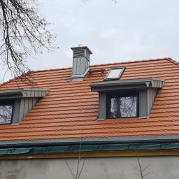 AMJ SPERO SP. Z O.O. - Czerwony dach z dachówką ceramiczną, szare okna dachowe i komin na tle pochmurnego nieba. Widoczny fragment elewacji budynku i drzewa.