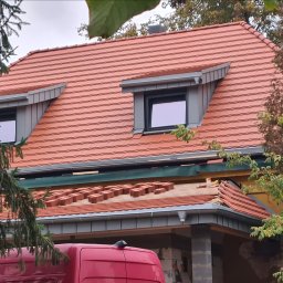 AMJ SPERO SP. Z O.O. - Częściowo pokryty dach z oknami dachowymi, widoczne ułożone dachówki w kolorze terakoty. Prace dekarskie w toku, fragment czerwonego samochodu na dole kadru.