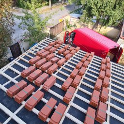 AMJ SPERO SP. Z O.O. - Przygotowane dachówki ceramiczne ułożone na drewnianej konstrukcji dachu, czerwony samochód dostawczy z otwartymi drzwiami w tle. Widok z góry na etap krycia.