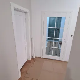 Jasne wnętrze po montażu białych drzwi wewnętrznych z szybami. Podłoga zabezpieczona kartonem. Widoczne dwie pary drzwi.