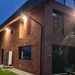AMJ SPERO SP. Z O.O. - Dwukondygnacyjny dom z cegły klinkierowej z dużymi oknami, oświetlony wieczornym światłem. Widoczne elementy dachu i otoczenie budynku.