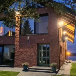 Nowoczesny dom z cegły klinkierowej oświetlony wieczorem. Duże okna, zielone drzwi wejściowe, zadbany trawnik i widok na jezioro w tle.