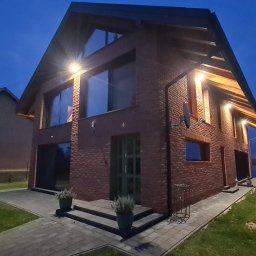 AMJ SPERO SP. Z O.O. - Dwupiętrowy dom z czerwonej cegły w nocnej scenerii. Widoczne duże okna, oświetlenie zewnętrzne i zadbany trawnik. Ujęcie z perspektywy ogrodu.