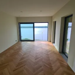 Jasne wnętrze z parkietem ułożonym w jodełkę, duże okno z widokiem na jezioro. Świeżo pomalowane ściany w odcieniach bieli i beżu, widoczne gniazdka elektryczne.