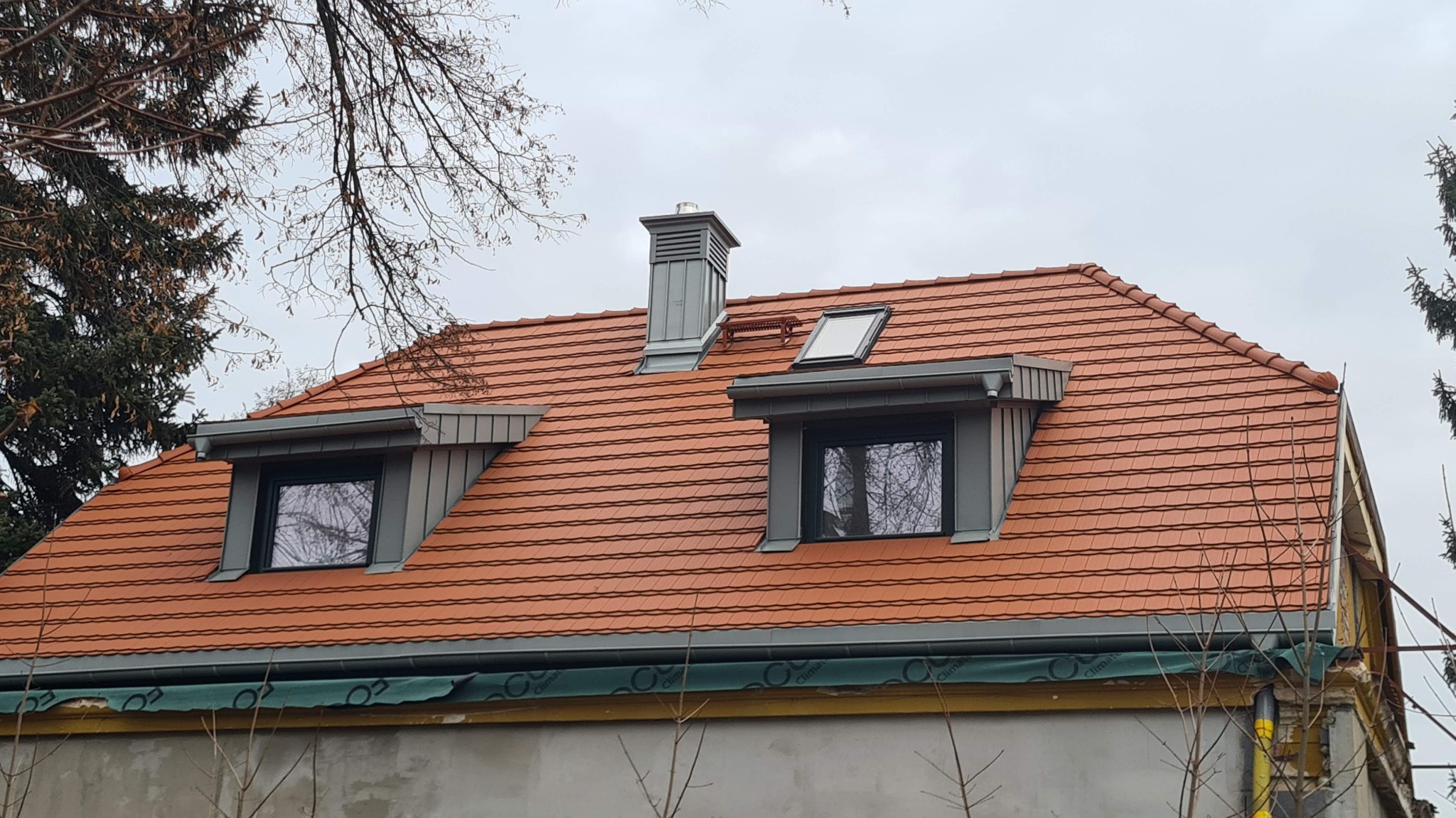 Czerwony dach z dachówką ceramiczną, szare okna dachowe i komin na tle pochmurnego nieba. Widoczny fragment elewacji budynku i drzewa.