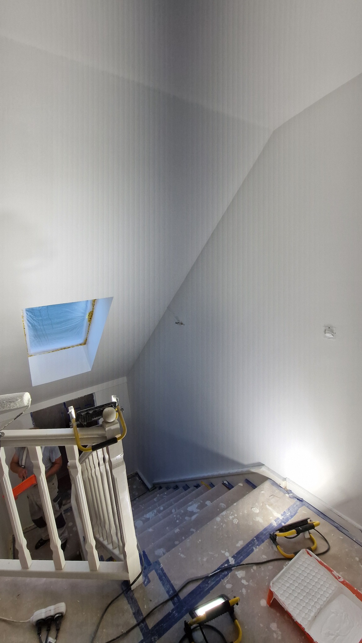 Świeżo pomalowane na biało ściany klatki schodowej z oknem dachowym, widok z góry. Na schodach widoczne zabezpieczenia i narzędzia malarskie.