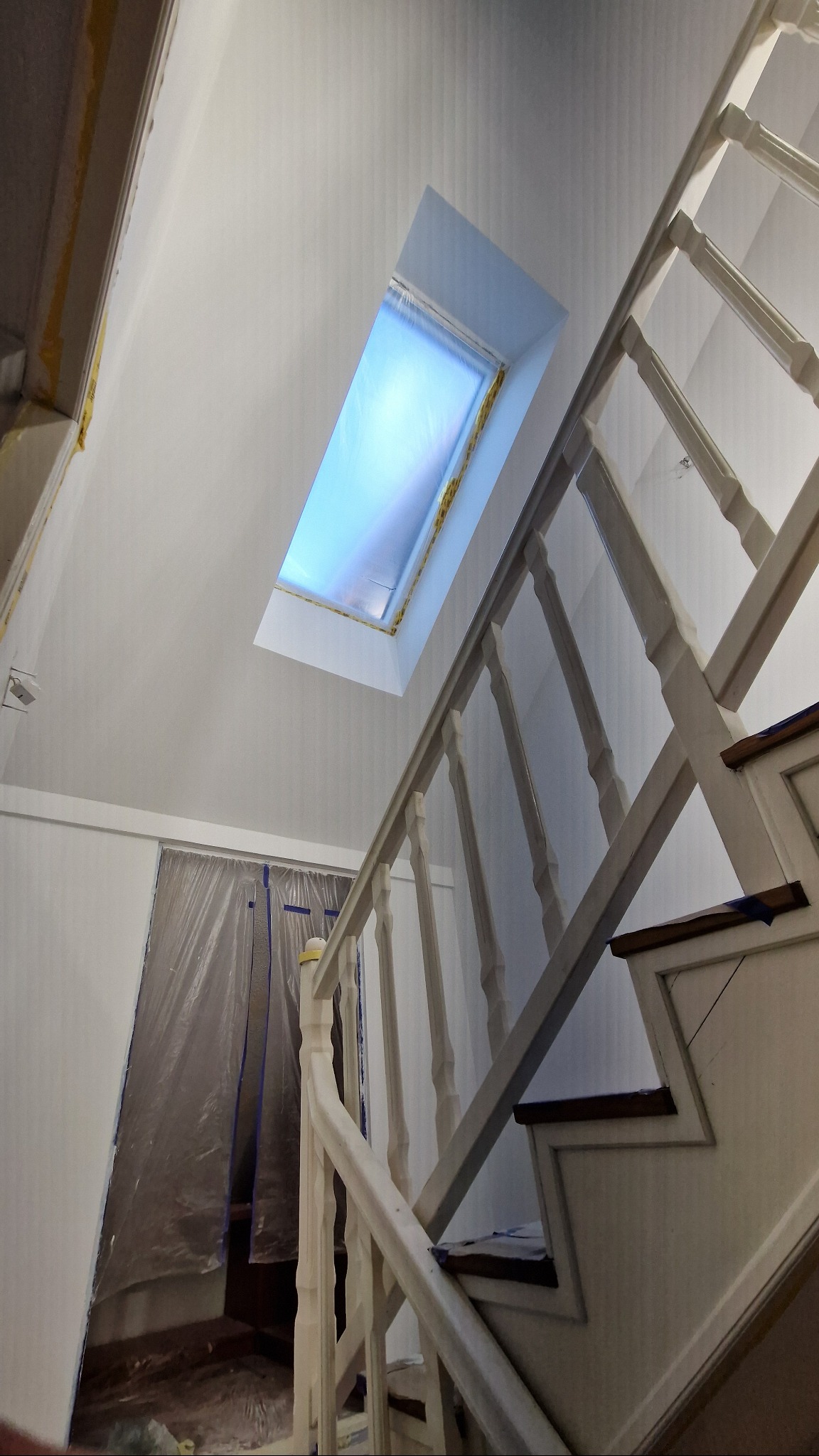 Białe schody z drewnianą balustradą i okno dachowe zabezpieczone folią malarską. Świeżo pomalowane ściany i sufit. Remont klatki schodowej.