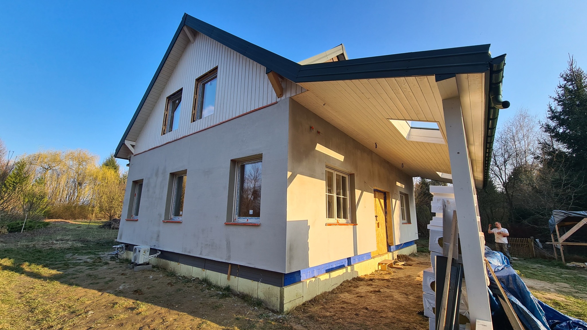 Dom jednorodzinny w trakcie prac wykończeniowych elewacji, widoczne okna, ocieplenie fundamentów i fragment dachu. Jasne, wiosenne światło.