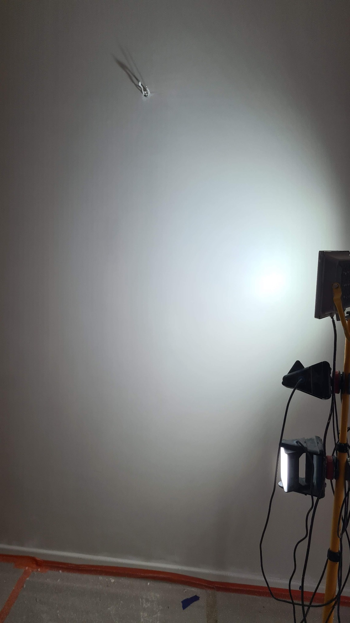 Świeżo pomalowana ściana oświetlona lampami budowlanymi, widoczne zabezpieczenia podłogi i wystający kabel ze ściany. Ściana w jasnym odcieniu szarości.