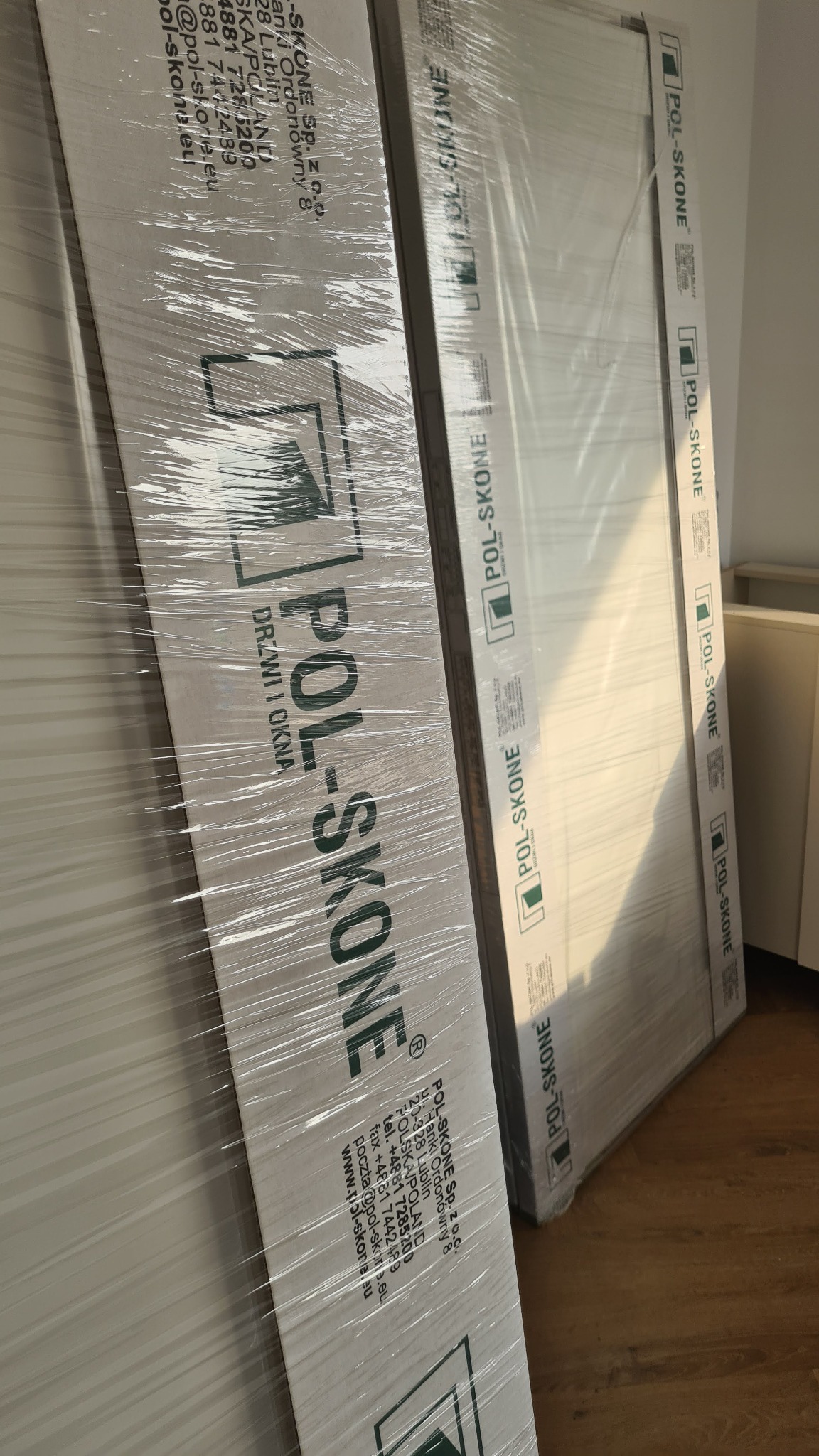 Dwa nowe, zapakowane drzwi marki Pol-Skone, ustawione obok siebie, gotowe do montażu. Widoczne logo producenta i adres firmy. Folia ochronna na powierzchni drzwi.