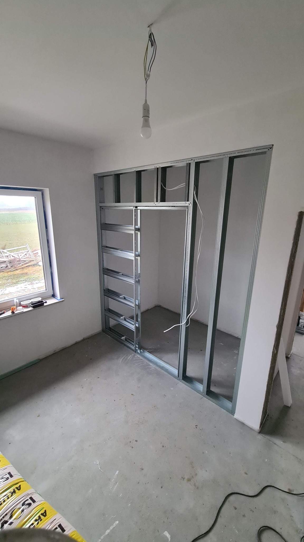 Wnętrze w trakcie prac: widok na metalową konstrukcję ścianki działowej z płyty karton-gips, z oknem w tle i wiszącą żarówką. Etap wznoszenia ścian działowych.