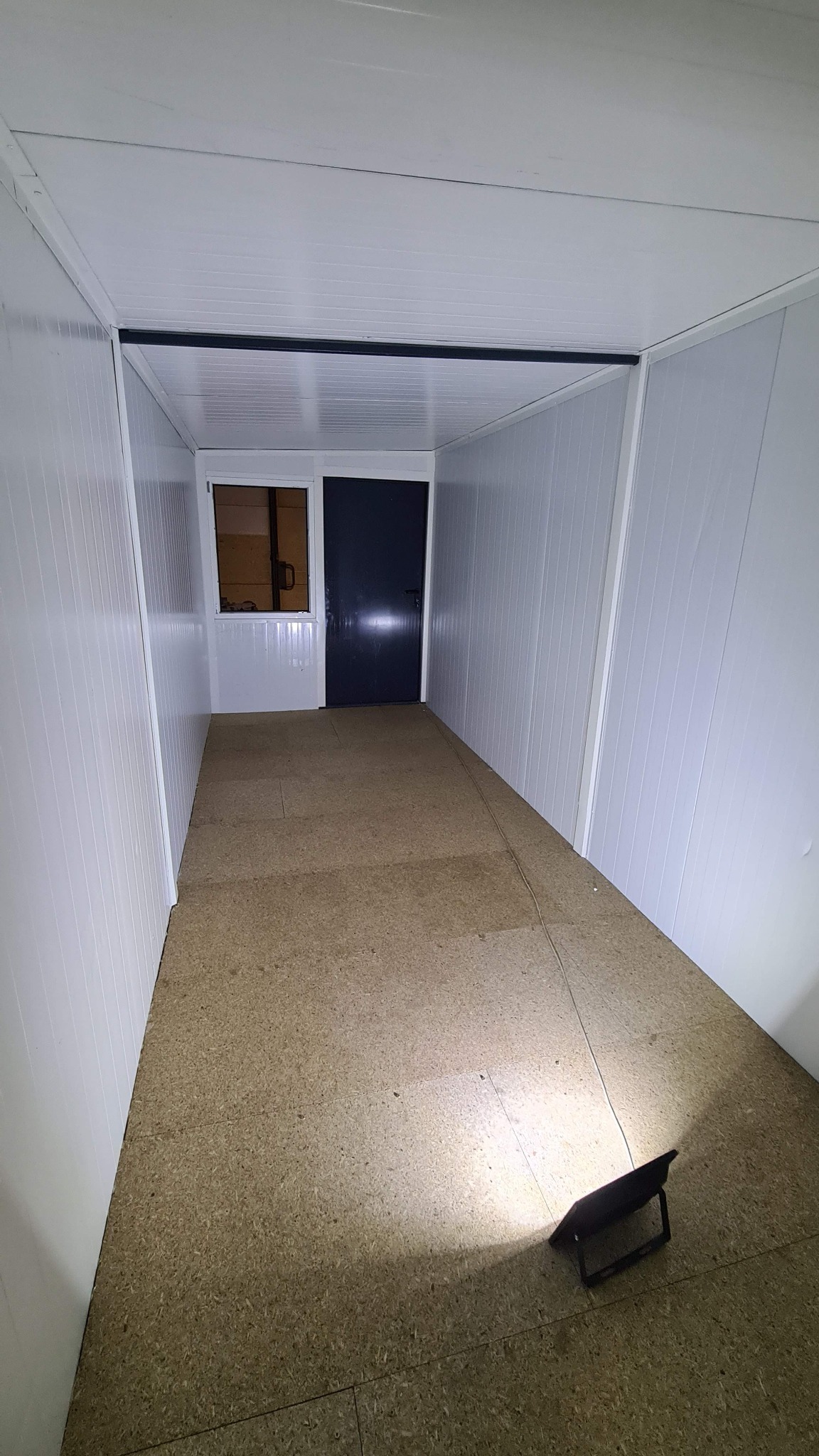 Wnętrze kontenera biurowego z panelami ściennymi, podłogą z płyt OSB i oknem. Widoczna lampa robocza. Surowe wykończenie, przygotowanie do dalszych prac.