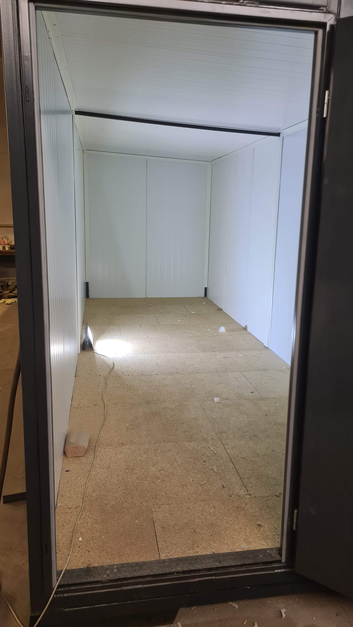Wnętrze chłodni z panelami izolacyjnymi i podłogą z płyt. Widoczny otwarty, ciemny futryna drzwi. Na podłodze resztki materiału.
