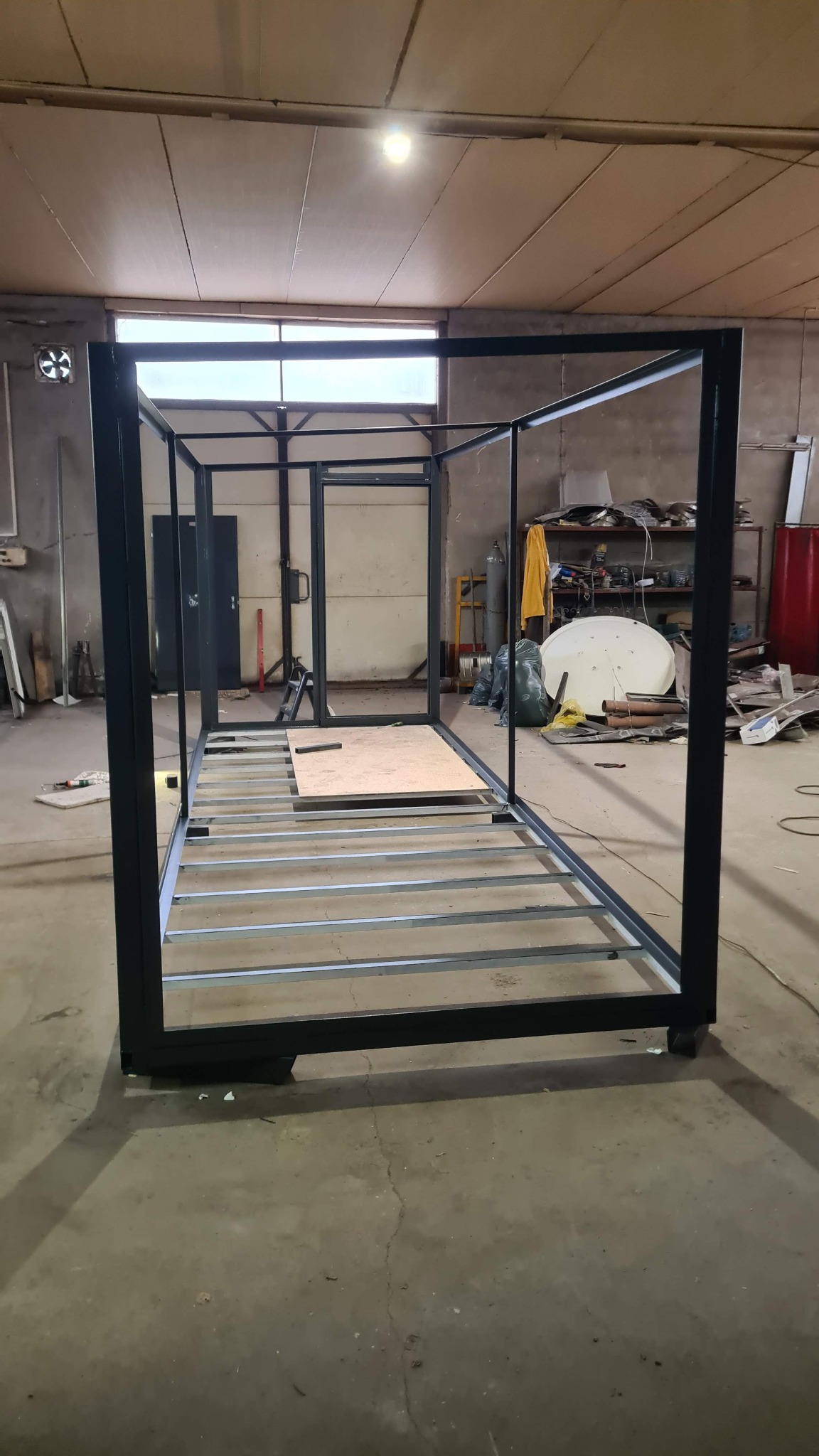 Metalowa konstrukcja szkieletowa w warsztacie, z widocznymi elementami podłogi i ramą, w trakcie montażu. Widoczne narzędzia i materiały w tle.