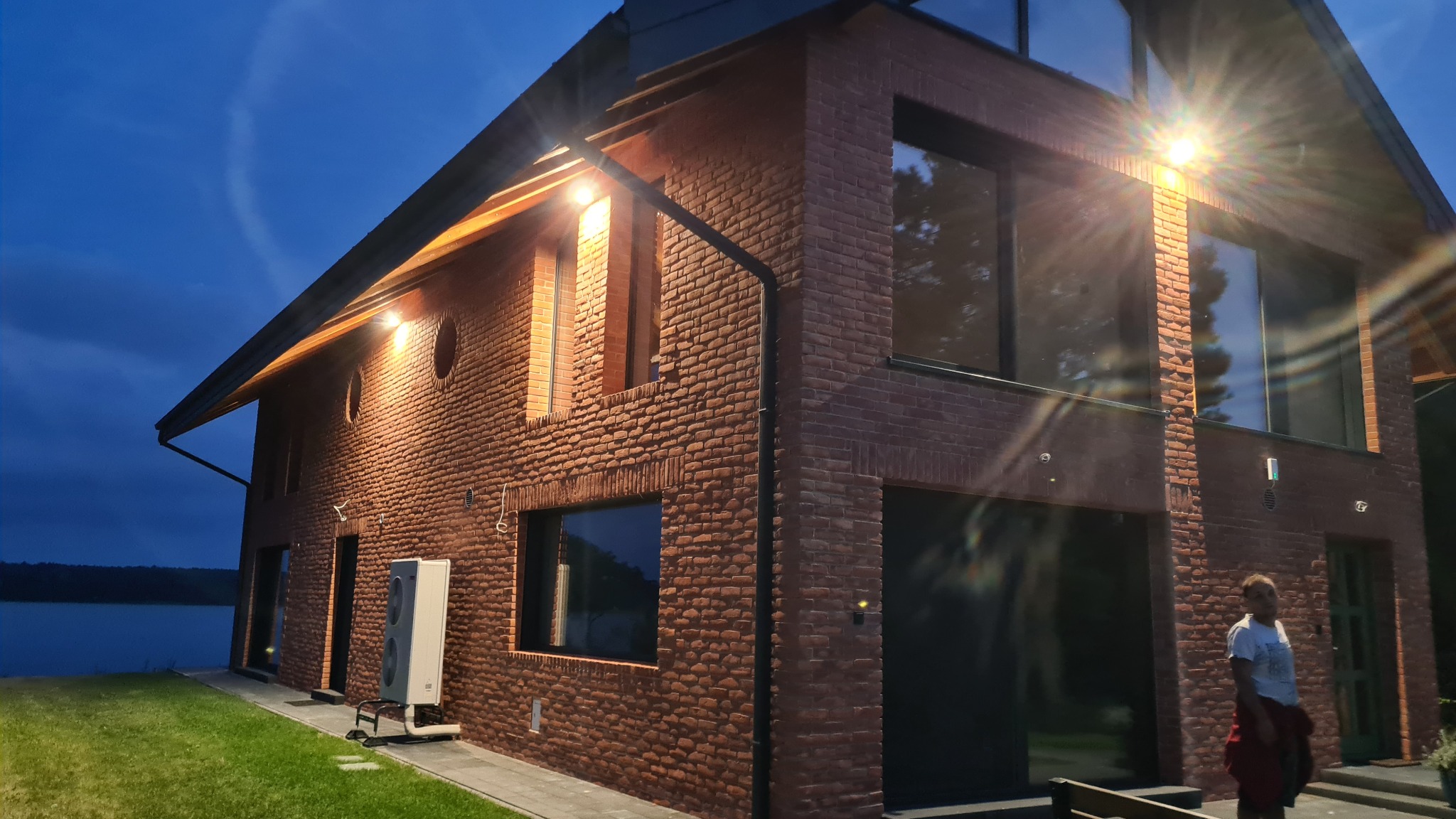 Dwukondygnacyjny dom z cegły klinkierowej z dużymi oknami, oświetlony wieczornym światłem. Widoczne elementy dachu i otoczenie budynku.