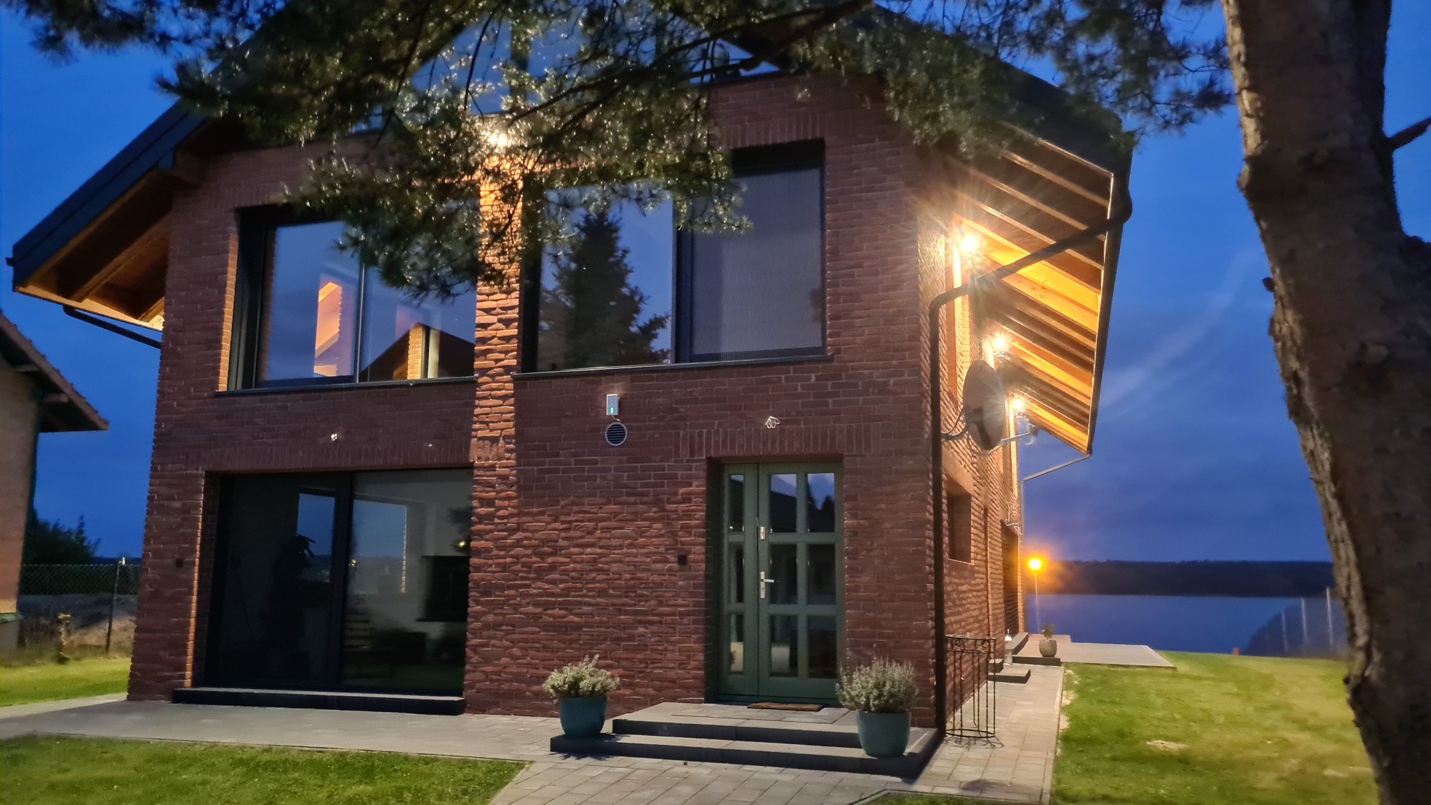 Nowoczesny dom z cegły klinkierowej oświetlony wieczorem. Duże okna, zielone drzwi wejściowe, zadbany trawnik i widok na jezioro w tle.