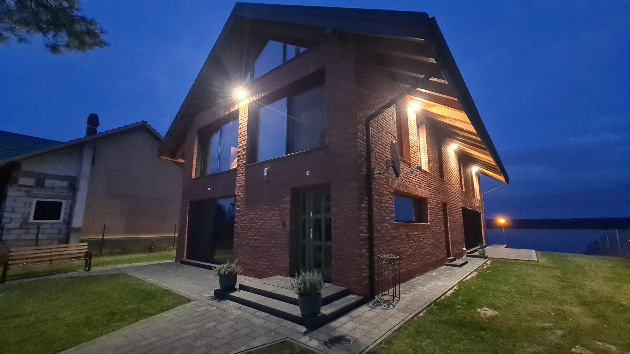 Dwupiętrowy dom z czerwonej cegły w nocnej scenerii. Widoczne duże okna, oświetlenie zewnętrzne i zadbany trawnik. Ujęcie z perspektywy ogrodu.