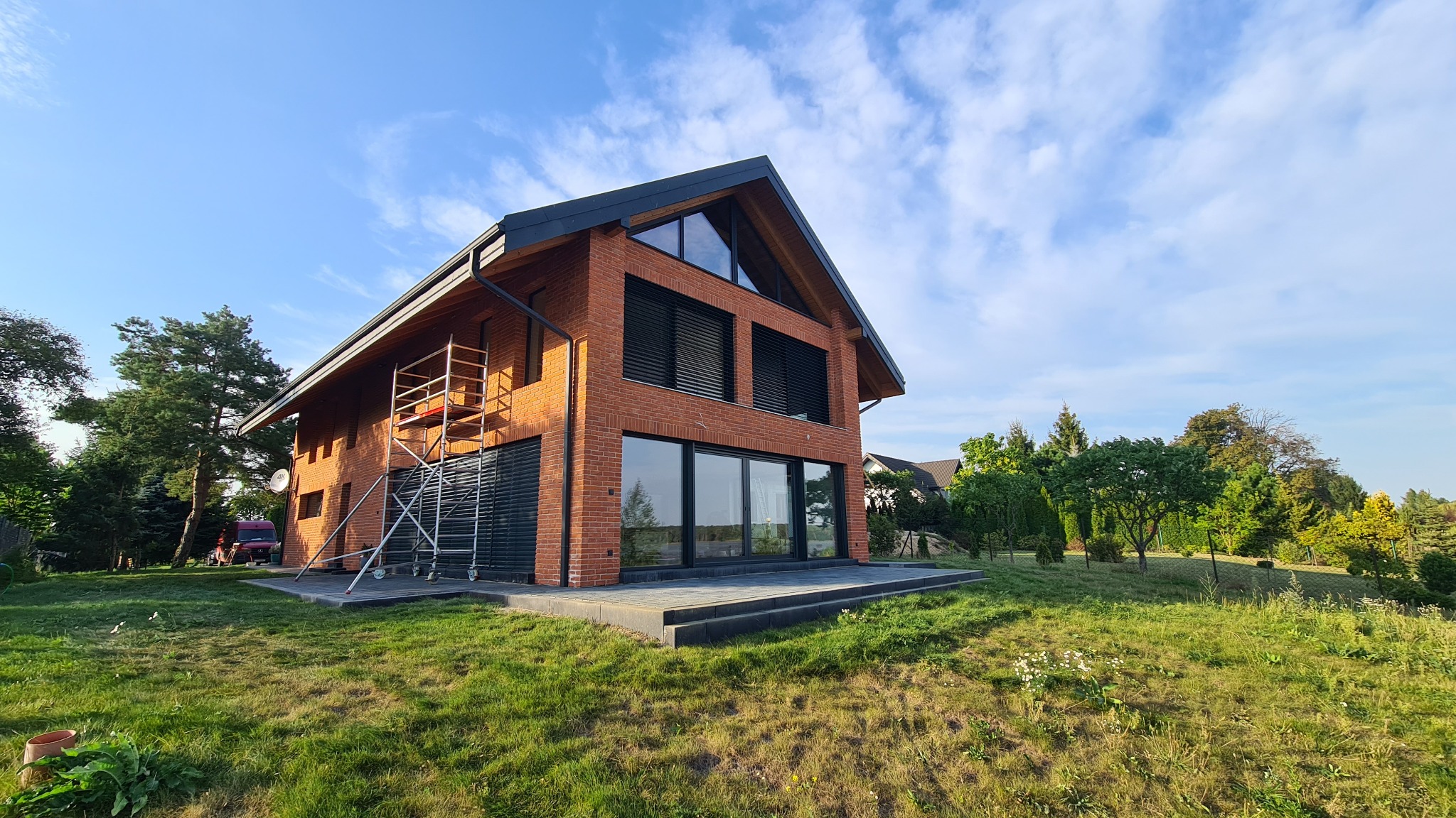 Nowoczesny dom z czerwonej cegły z dużymi oknami i czarnymi żaluzjami, widok z ogrodu. Na bocznej ścianie rusztowanie. Uwagę przykuwa wykończenie elewacji.