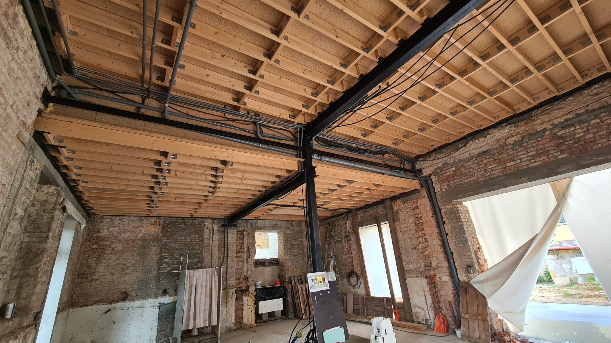 Wnętrze w trakcie remontu: drewniany strop z instalacją, surowe ceglane ściany z oknami, widoczne elementy konstrukcyjne i instalacje. Charakterystyczny etap prac budowlanych.
