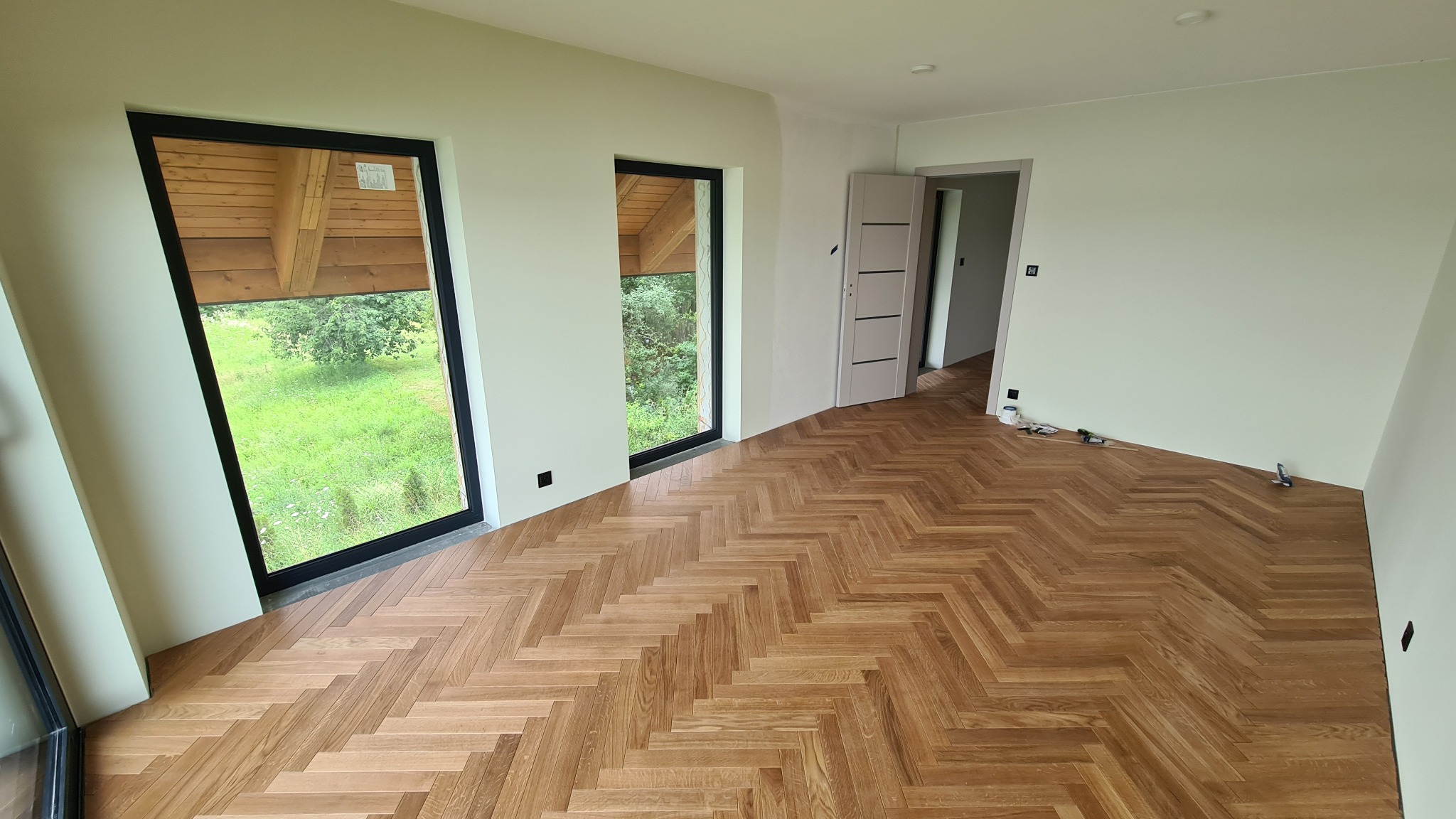 Jasne wnętrze z parkietem ułożonym we wzór jodełki. Czarne ramy okien z widokiem na zieleń. Otwarta, biała ściana z szarymi drzwiami wewnętrznymi w tle.